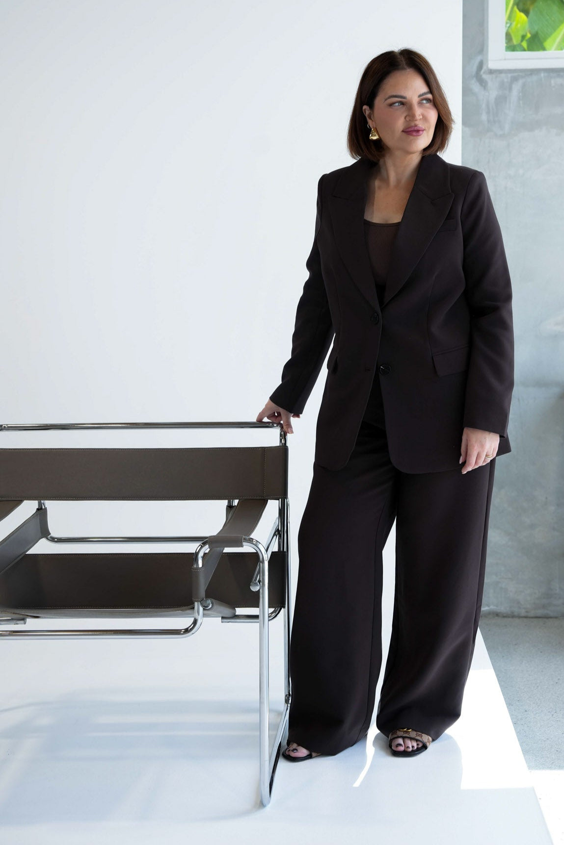 Woman in a black suit standing in a modern interior setting with a plant in the background.